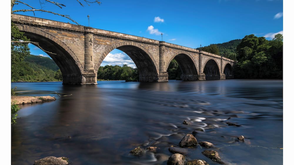 Eine Steinbrücke über einem Fluss mit klarem Wasser und Steinen im Vordergrund.
