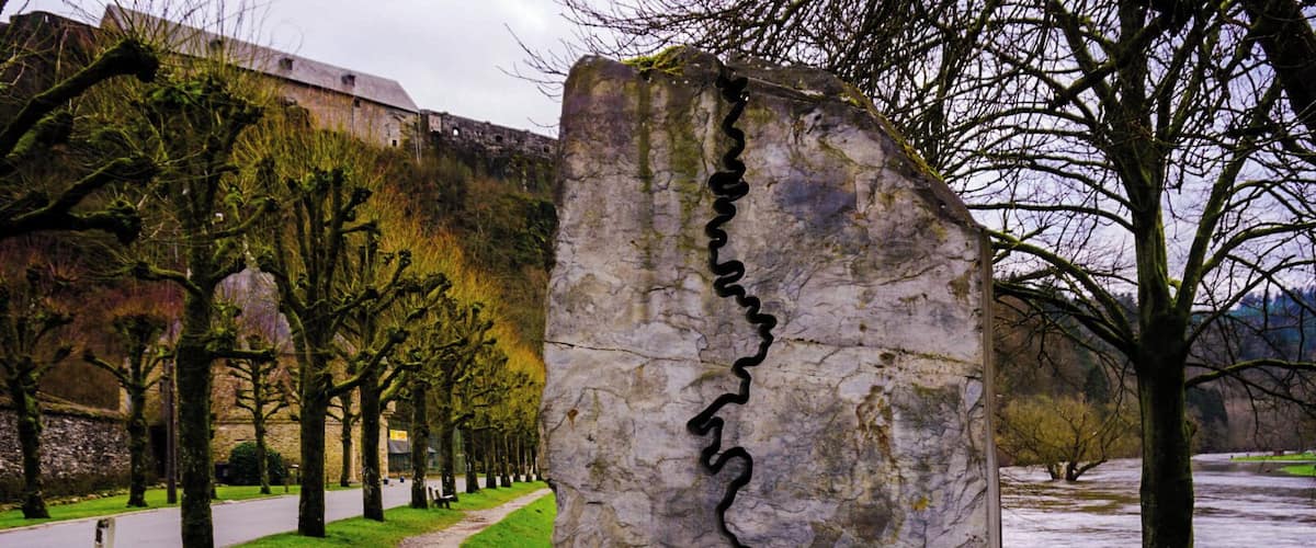 A stone monument with a crack running through it, surrounded by a pathway, trees, and a body of water.