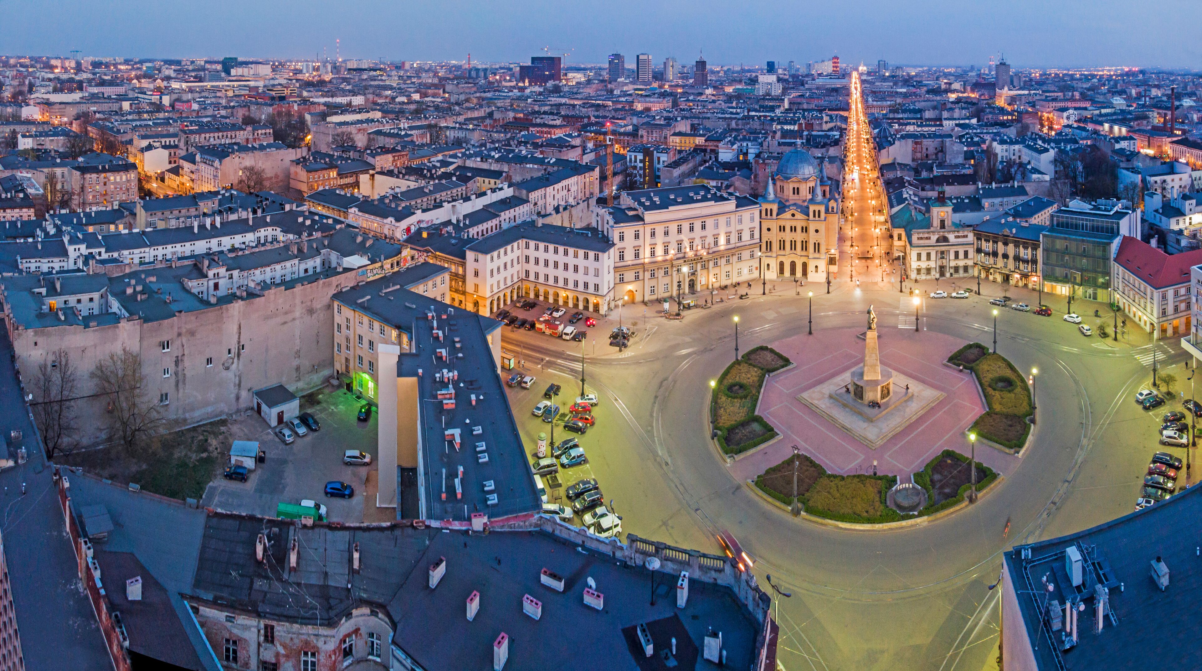 A cityscape at dusk with a prominent roundabout, historic buildings, and a central monument.