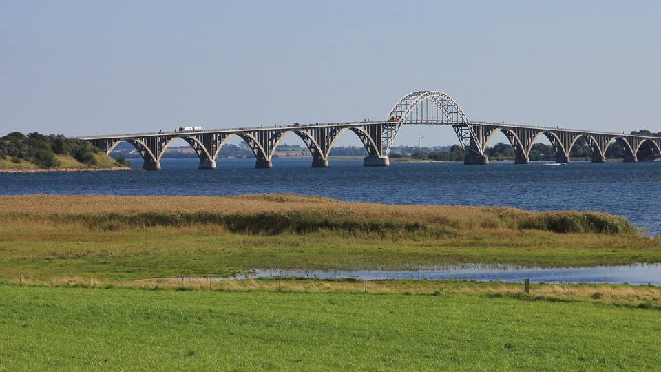 A large steel bridge with a distinctive arch over a river, surrounded by grassy fields and trees.