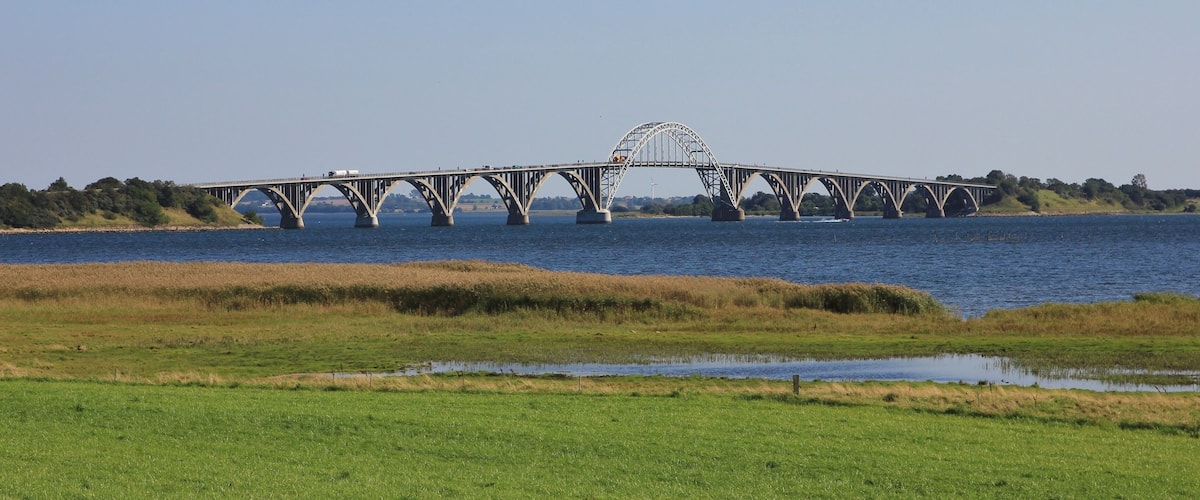 A large steel bridge with a distinctive arch over a river, surrounded by grassy fields and trees.