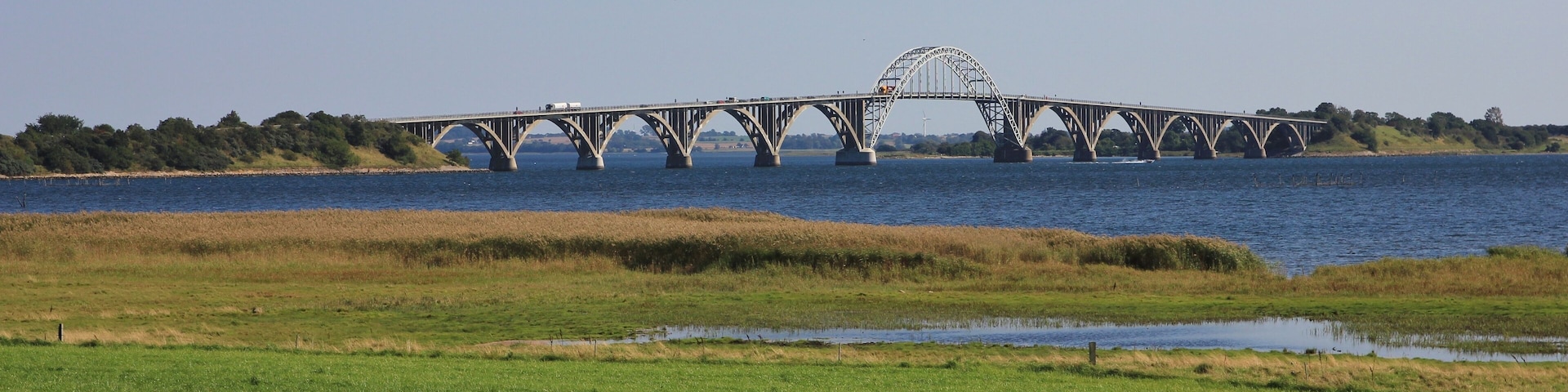 A large steel bridge with a distinctive arch over a river, surrounded by grassy fields and trees.