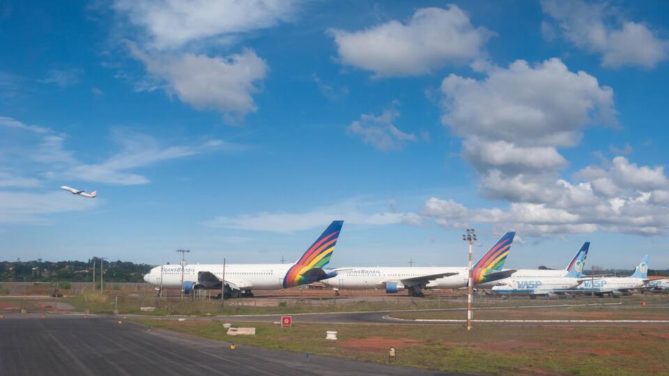 An airport with three airplanes parked on the tarmac, a clear blue sky, and a small aircraft flying in the distance.