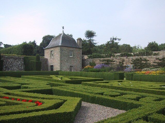 A stone building with a dome roof surrounded by a formal garden with geometric hedges and flower beds.