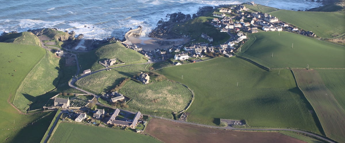 An aerial view of a coastal village with green fields, a golf course, and a rocky shoreline.