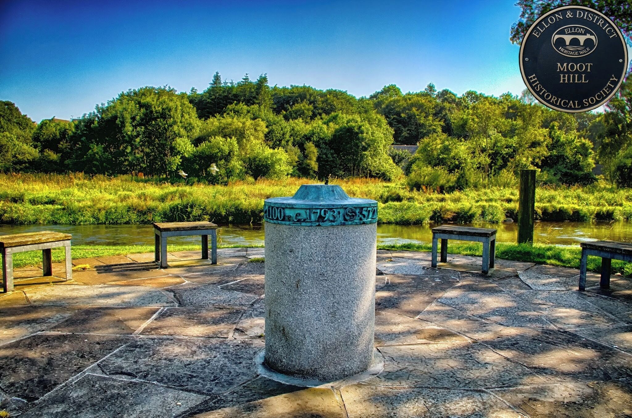 Monumento de piedra con inscripciones, rodeado de bancos de piedra y un río.