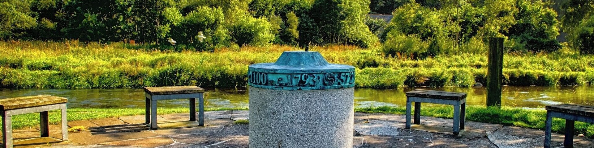 Monumento de piedra con inscripciones, rodeado de bancos de piedra y un río.