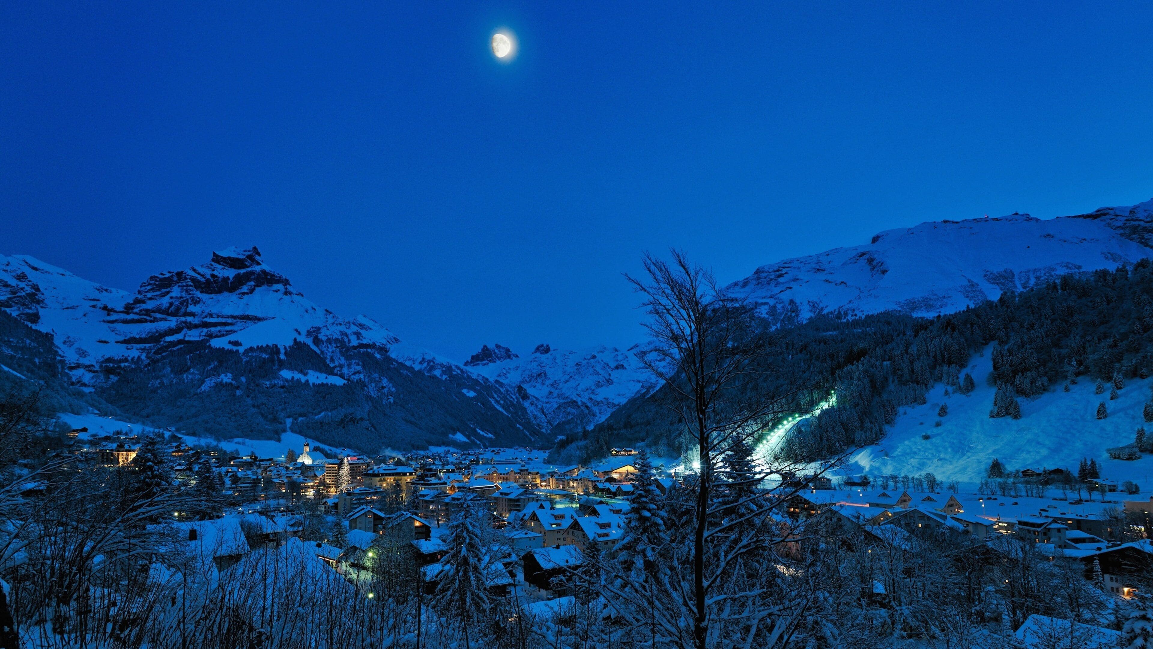 A snow-covered mountain village at night with a crescent moon in the sky.