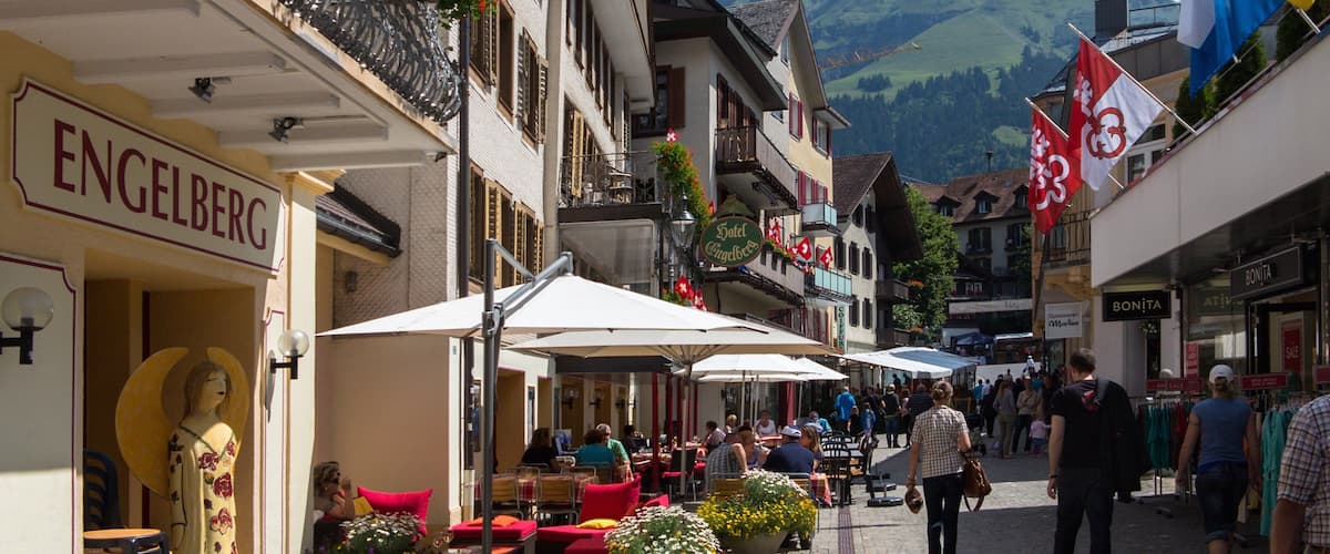 A bustling street with outdoor cafes, a statue, and a mountain backdrop.