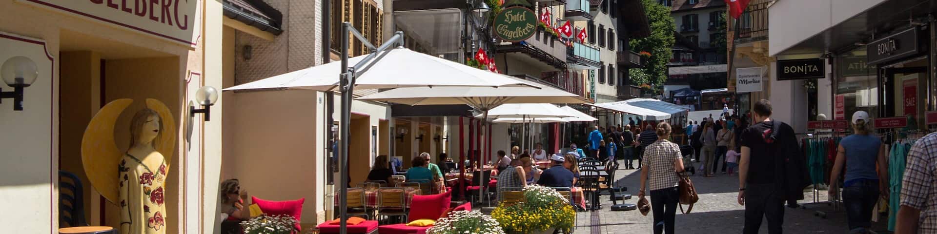 A bustling street with outdoor cafes, a statue, and a mountain backdrop.