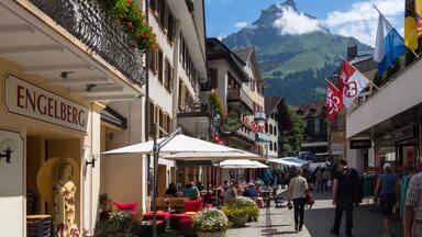 Una calle animada con cafés al aire libre, una estatua y montañas al fondo.