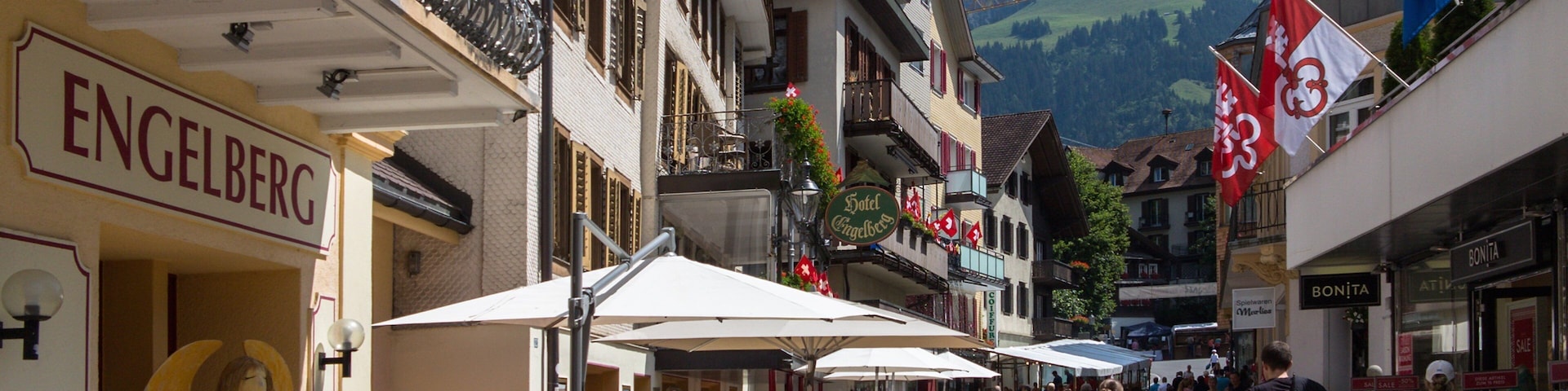 Una calle animada con cafés al aire libre, una estatua y montañas al fondo.