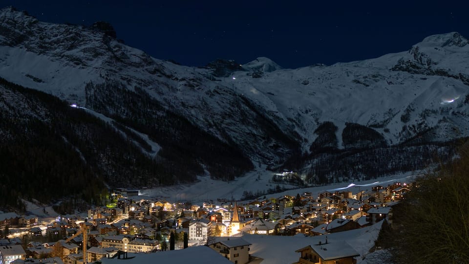 A snow-covered mountain town at night with illuminated buildings and a church spire.