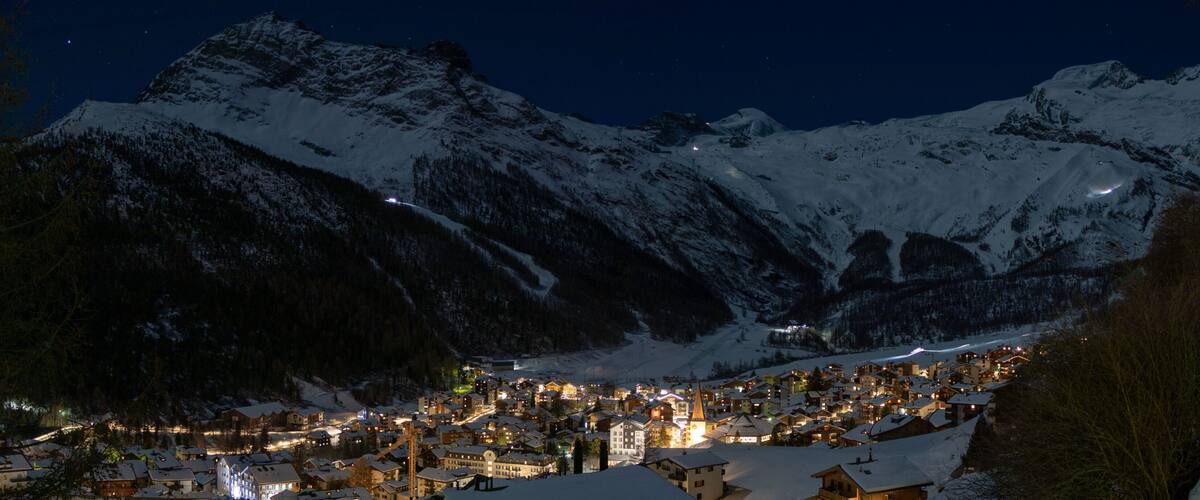 A snow-covered mountain town at night with illuminated buildings and a church spire.
