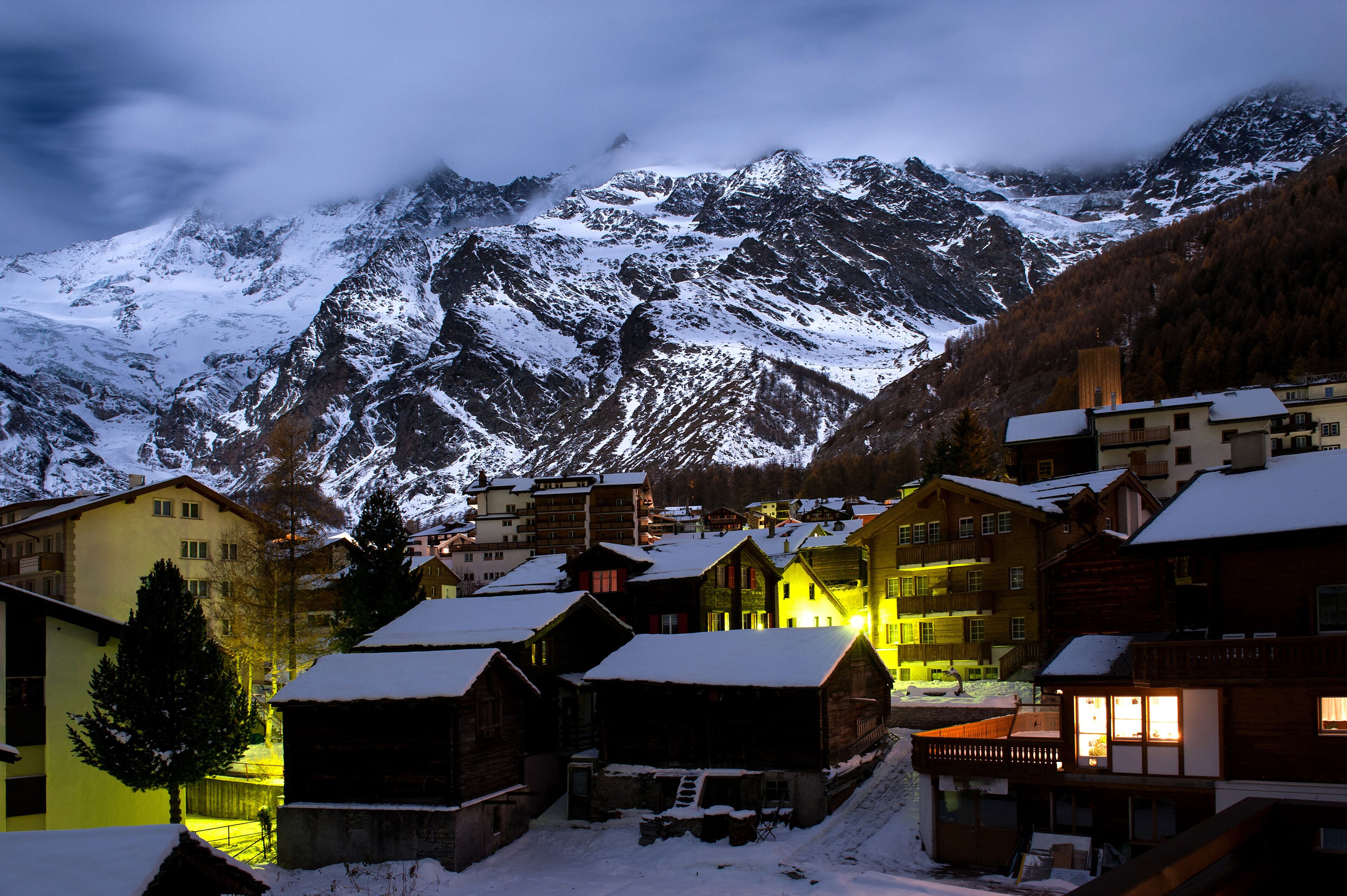 Panoramic view of the Swiss mountain resort of Saas-Fee at twilight, nestled in a valley beneath towering, snow-covered, 4,000-meter peaks.