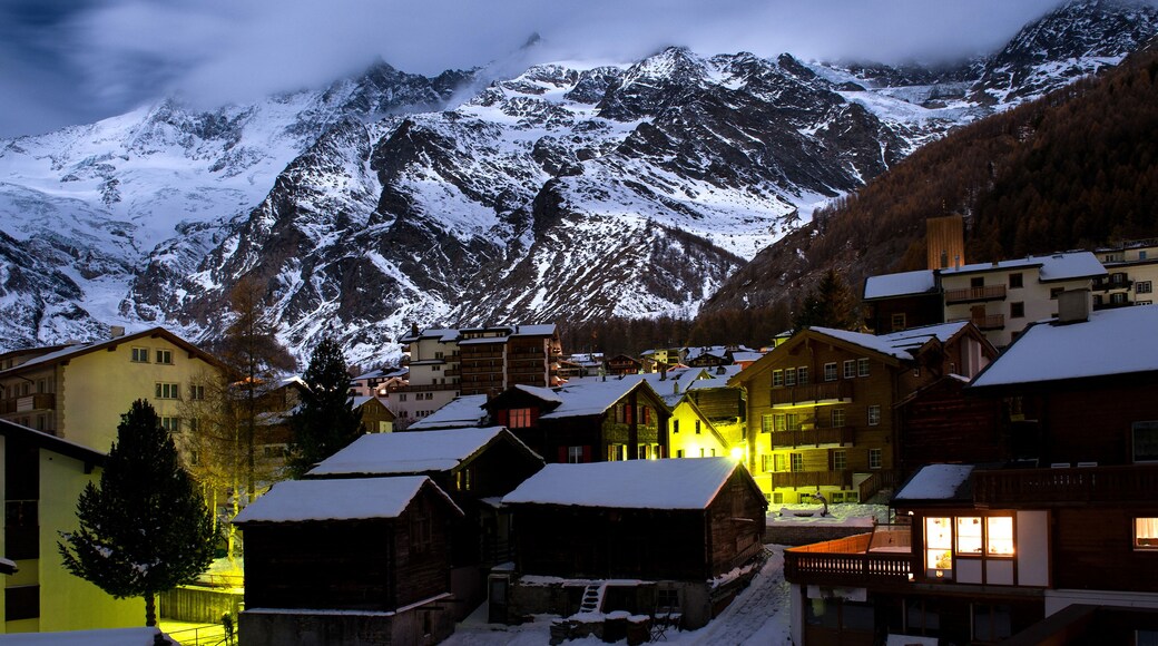 Panoramic view of the Swiss mountain resort of Saas-Fee at twilight, nestled in a valley beneath towering, snow-covered, 4,000-meter peaks.