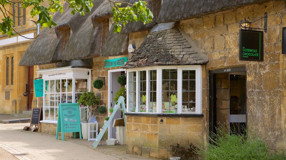 A traditional stone building with a thatched roof, a sign for 'The Cotswold Chocolate Company', and a shop named 'Whatever the Weather'.