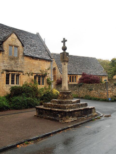 A stone church with a cross atop a pedestal, surrounded by stone walls and vegetation.