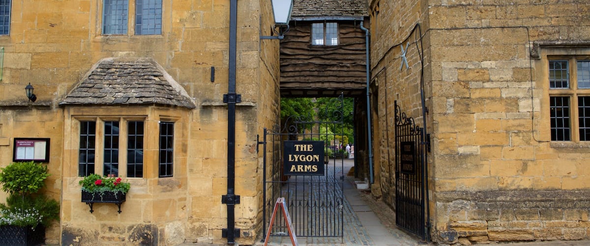 A narrow street with stone buildings, a sign for 'The Lygon Arms' pub, and a small garden with flowers.