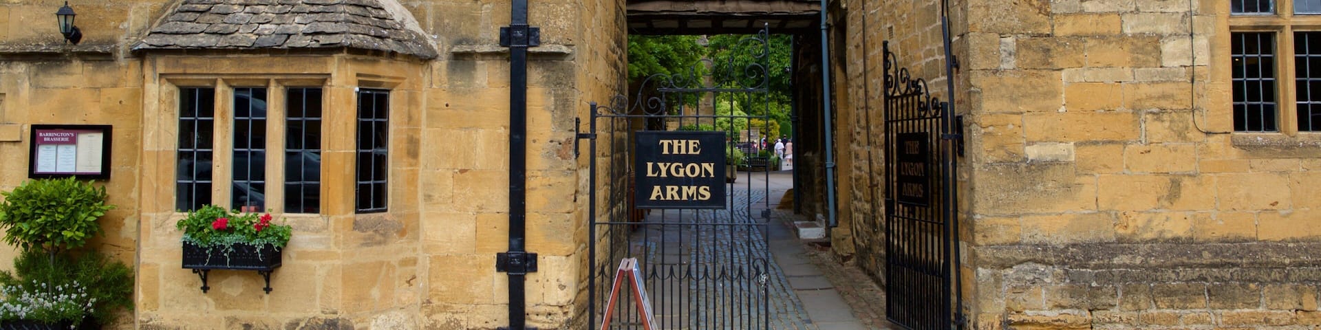 A narrow street with stone buildings, a sign for 'The Lygon Arms' pub, and a small garden with flowers.