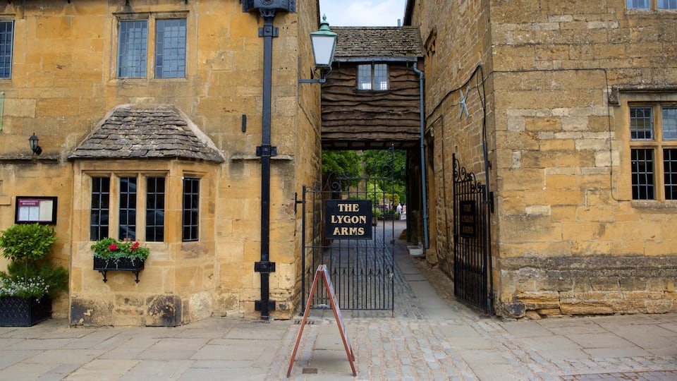 A narrow street with stone buildings, a sign for 'The Lygon Arms' pub, and a small garden with flowers.