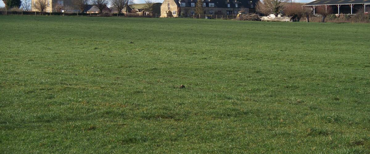 A large green field with a stone building and a farmhouse in the background.