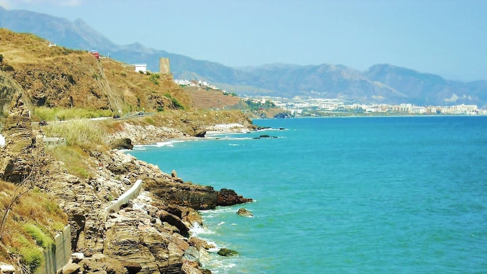 A coastal landscape with rocky cliffs, a clear blue sea, and distant mountains.