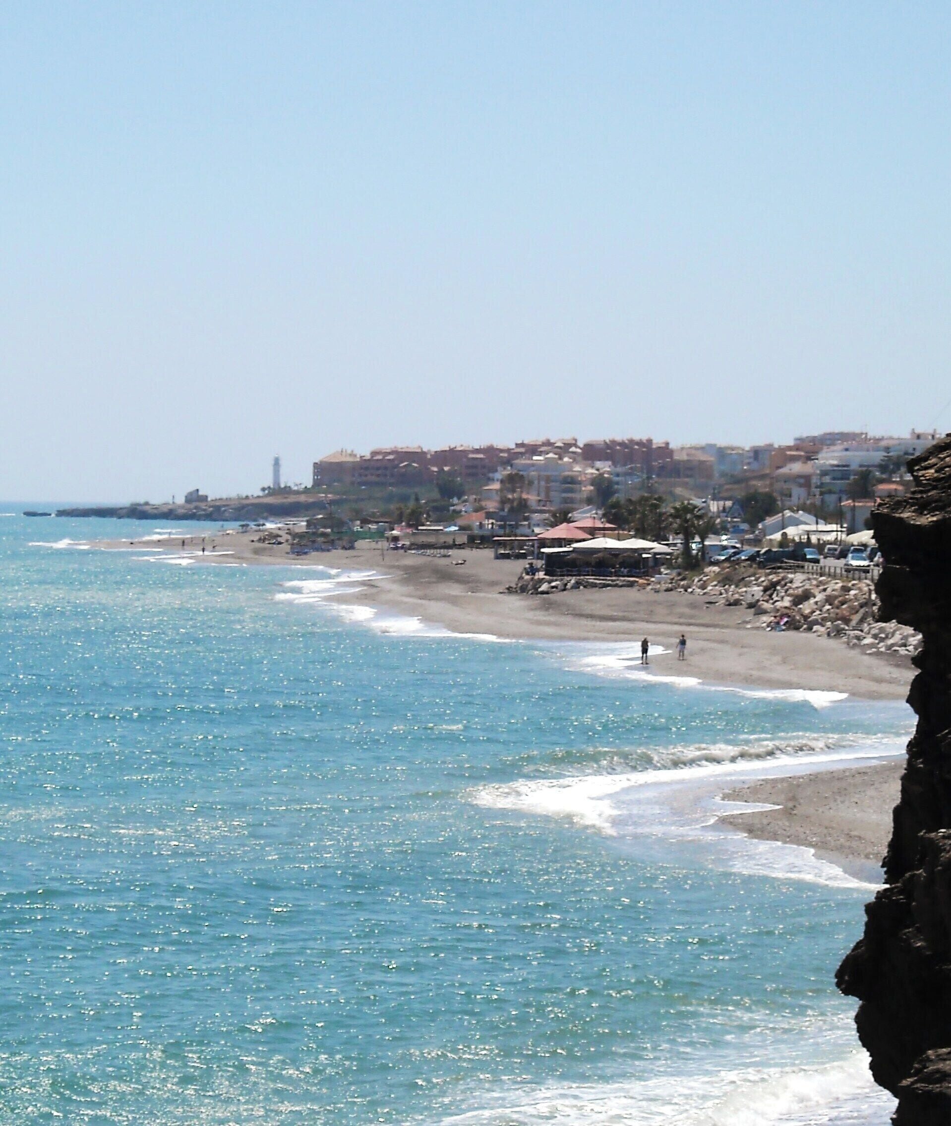 A coastal beach with clear blue water, rocky cliffs, and buildings in the background.