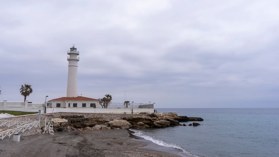 A coastal lighthouse with a white tower, a stone building with a red roof, and a rocky shoreline.