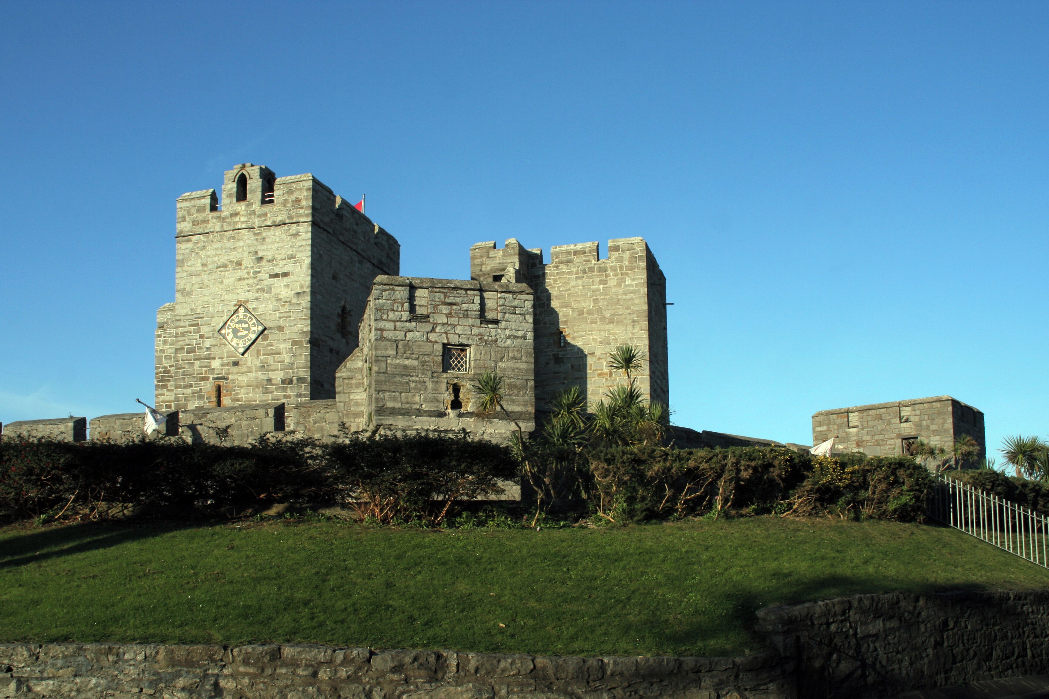A historic stone castle with battlements and a clock tower, surrounded by a well-maintained lawn and a stone wall.