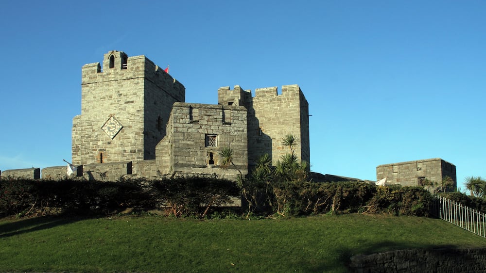 Eine historische Steinburg mit Zinnen und einem Glockenturm, umgeben von einer gepflegten Rasenfläche und einer Steinmauer.