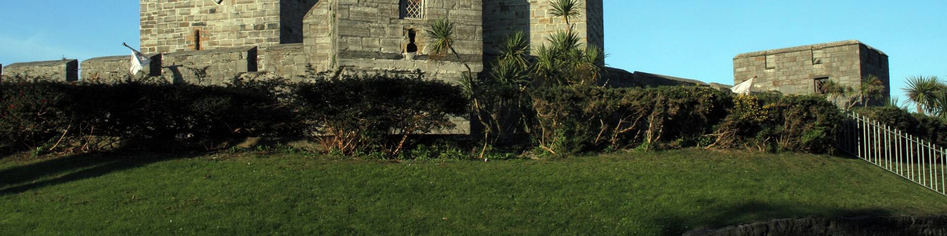 A historic stone castle with battlements and a clock tower, surrounded by a well-maintained lawn and a stone wall.
