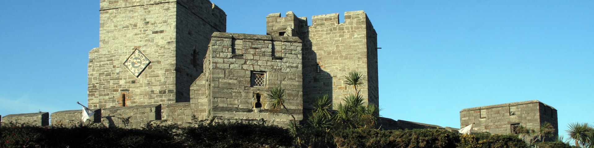 Un castillo de piedra histórico con almenas y una torre del reloj, rodeado de un césped bien cuidado y un muro de piedra.