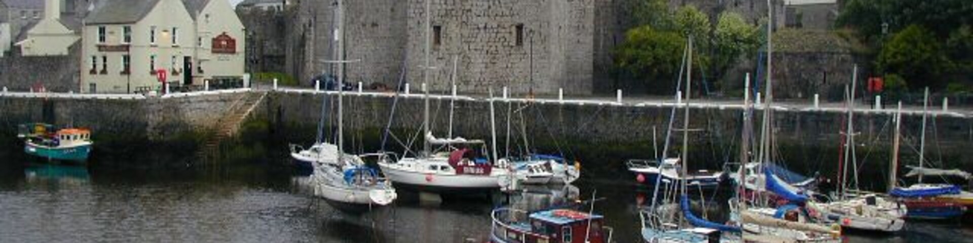 Un puerto con varias embarcaciones amarradas, un castillo al fondo y edificios a lo largo de la costa.