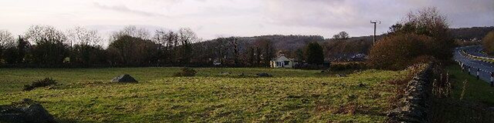 Paisaje rural con un muro de piedra, un campo cubierto de hierba y un edificio pequeño a lo lejos.