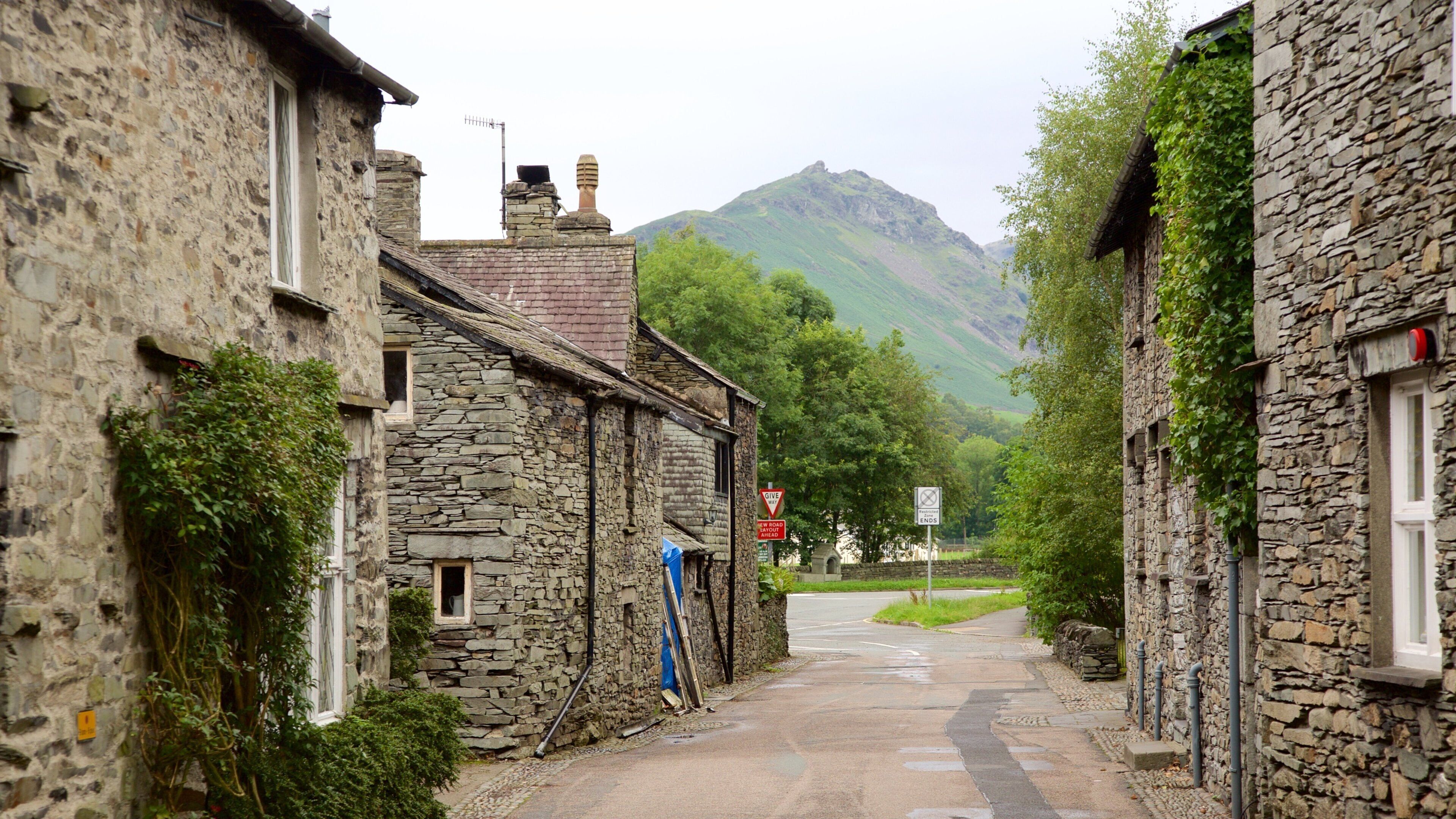 A narrow stone street flanked by traditional stone buildings, a 'Give Way' sign, and a 'New Road Ends' sign, with a mountain in the background.