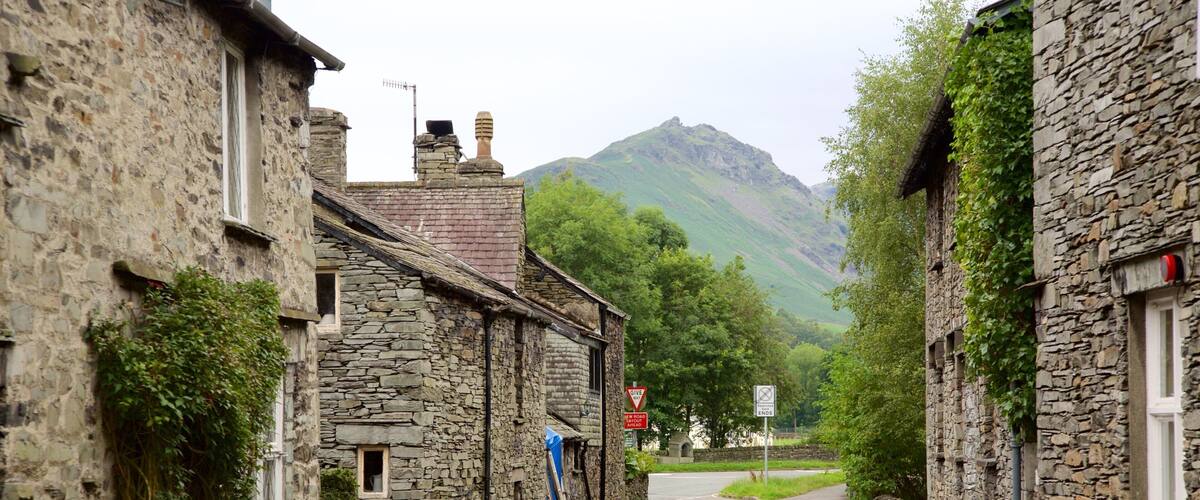 A narrow stone street flanked by traditional stone buildings, a 'Give Way' sign, and a 'New Road Ends' sign, with a mountain in the background.