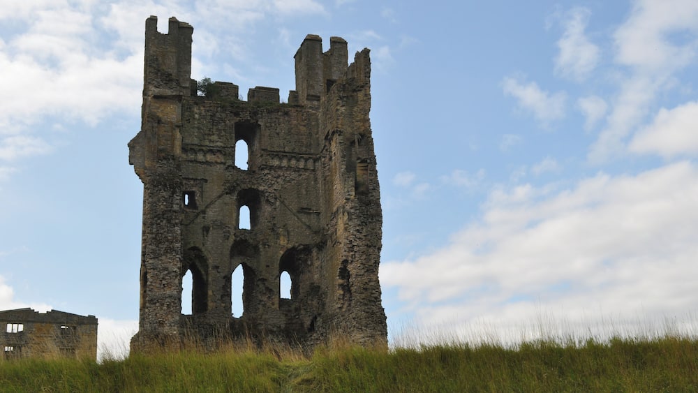 Une tour de pierre historique, dotée de nombreuses arcades et de créneaux, entourée de prairies verdoyantes et surplombée par un ciel partiellement nuageux.