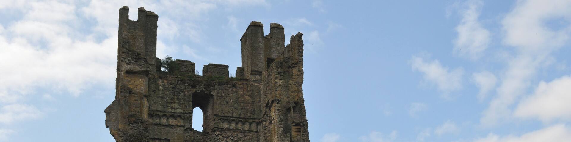 A historic stone tower with multiple arches and battlements, surrounded by grassy fields and a partly cloudy sky.
