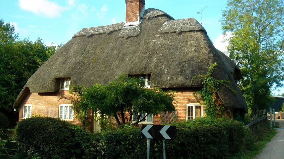 A thatched-roof house with a brick base, surrounded by greenery and a directional sign.