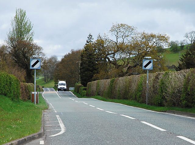 A rural road with a sharp bend to the right, flanked by hedges and trees.