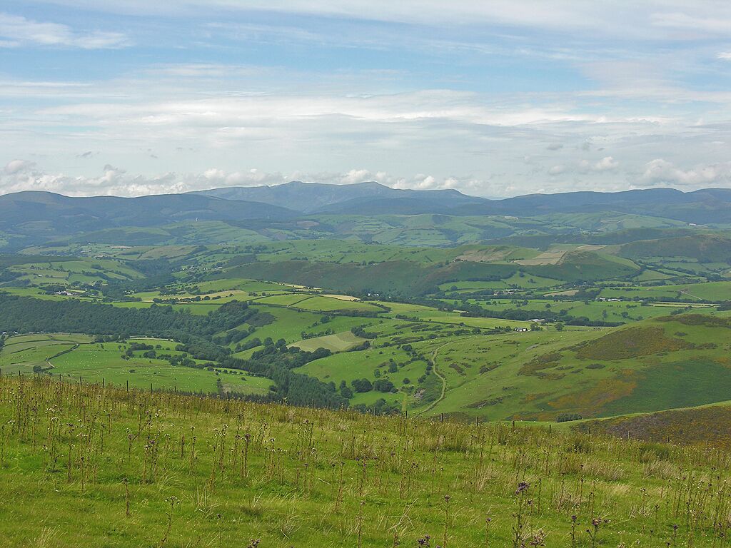 Rolling green hills, scattered fields, and distant mountains under a partly cloudy sky.