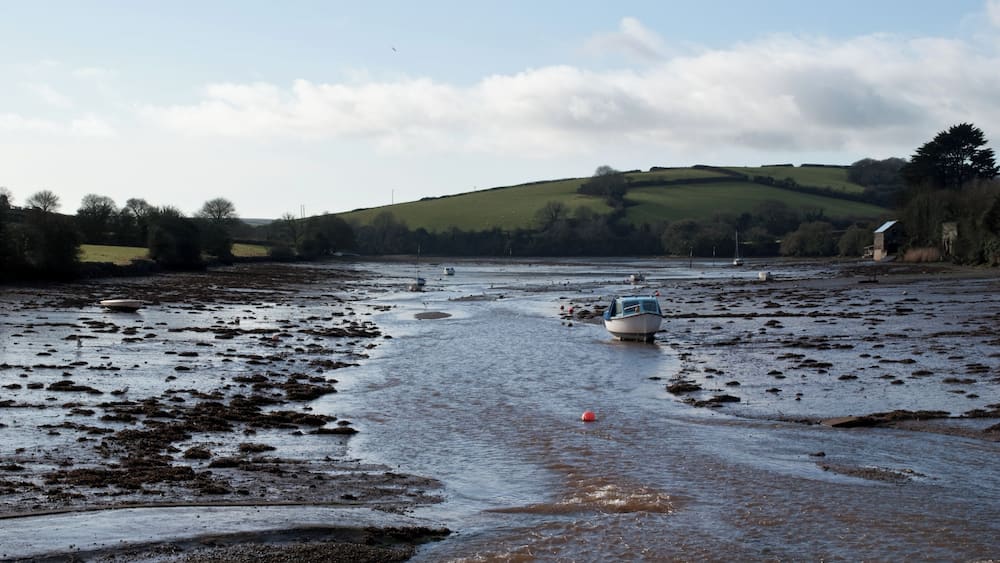 Une rivière avec des bateaux, des berges boueuses et une colline verdoyante en arrière-plan.