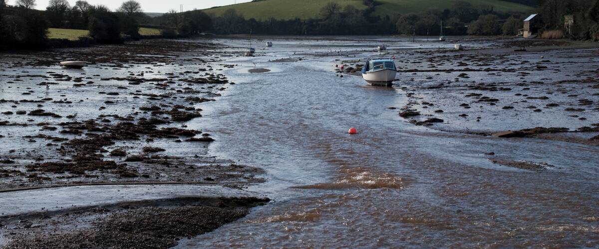 Une rivière avec des bateaux, des berges boueuses et une colline verdoyante en arrière-plan.