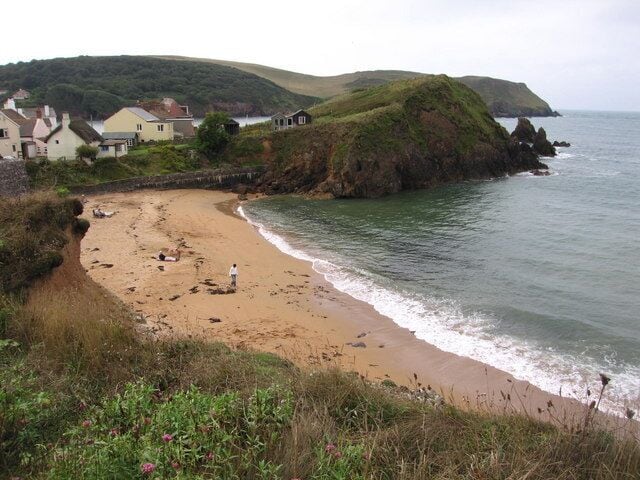 A coastal beach with houses on a hillside, a rocky outcrop, and a calm sea.