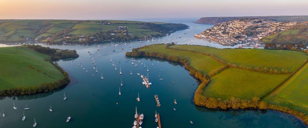 A coastal town with a harbor filled with sailboats, surrounded by green fields and hills.