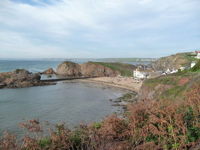 A coastal landscape with rocky outcrops, a sandy beach, and a few buildings on the hillside.