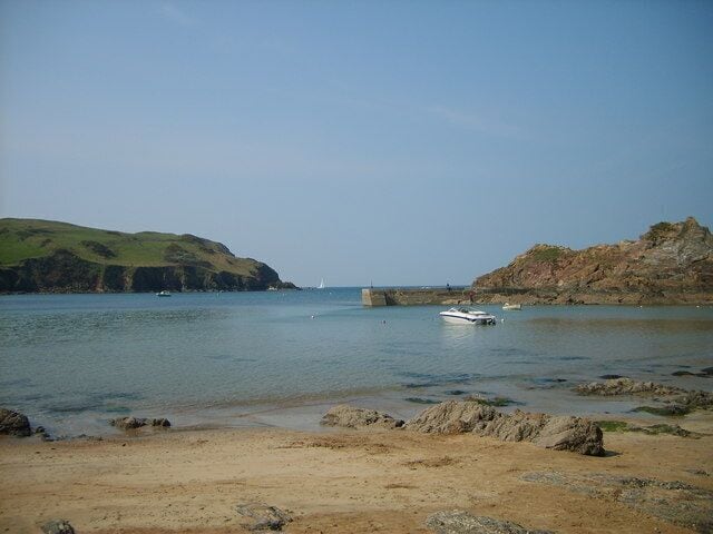 A coastal landscape with a sandy beach, calm water, rocky outcrops, and a small boat.