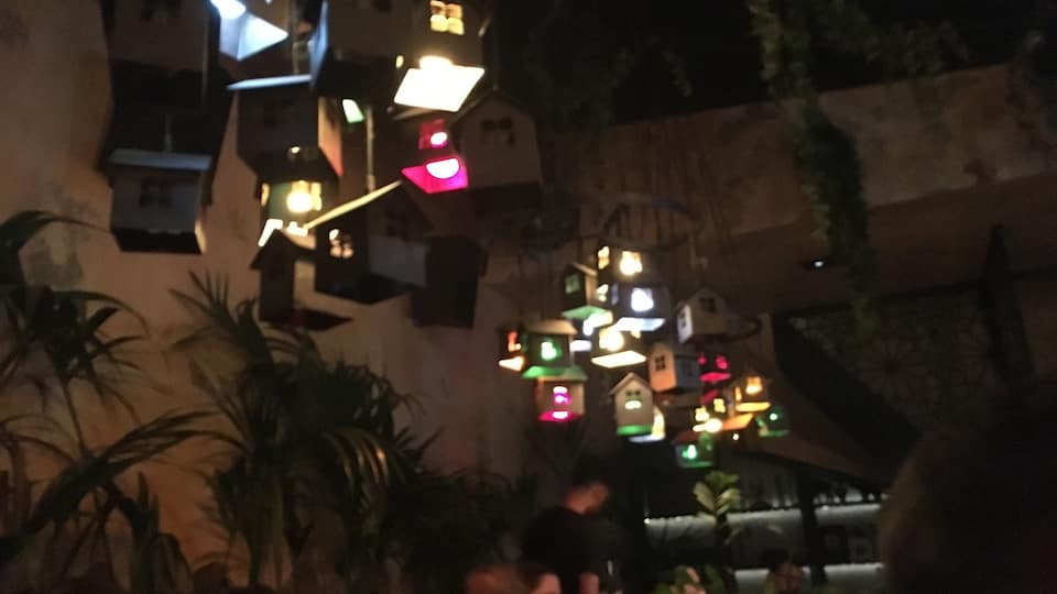 A row of illuminated birdhouses against a wall, with people seated at tables below.