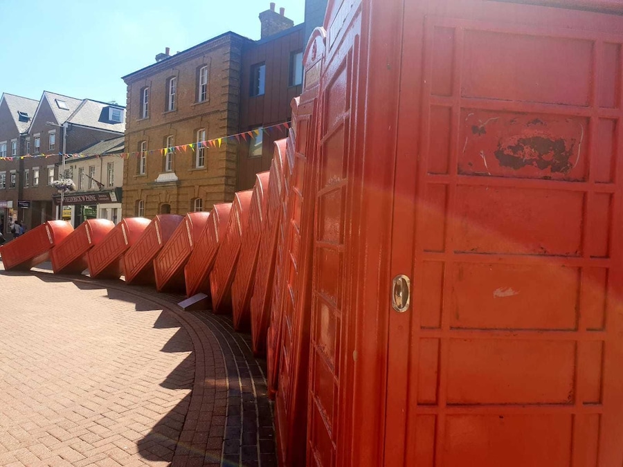 Uma fileira de portas vermelhas em uma praça pública com calçamento de tijolos e edifícios ao fundo.